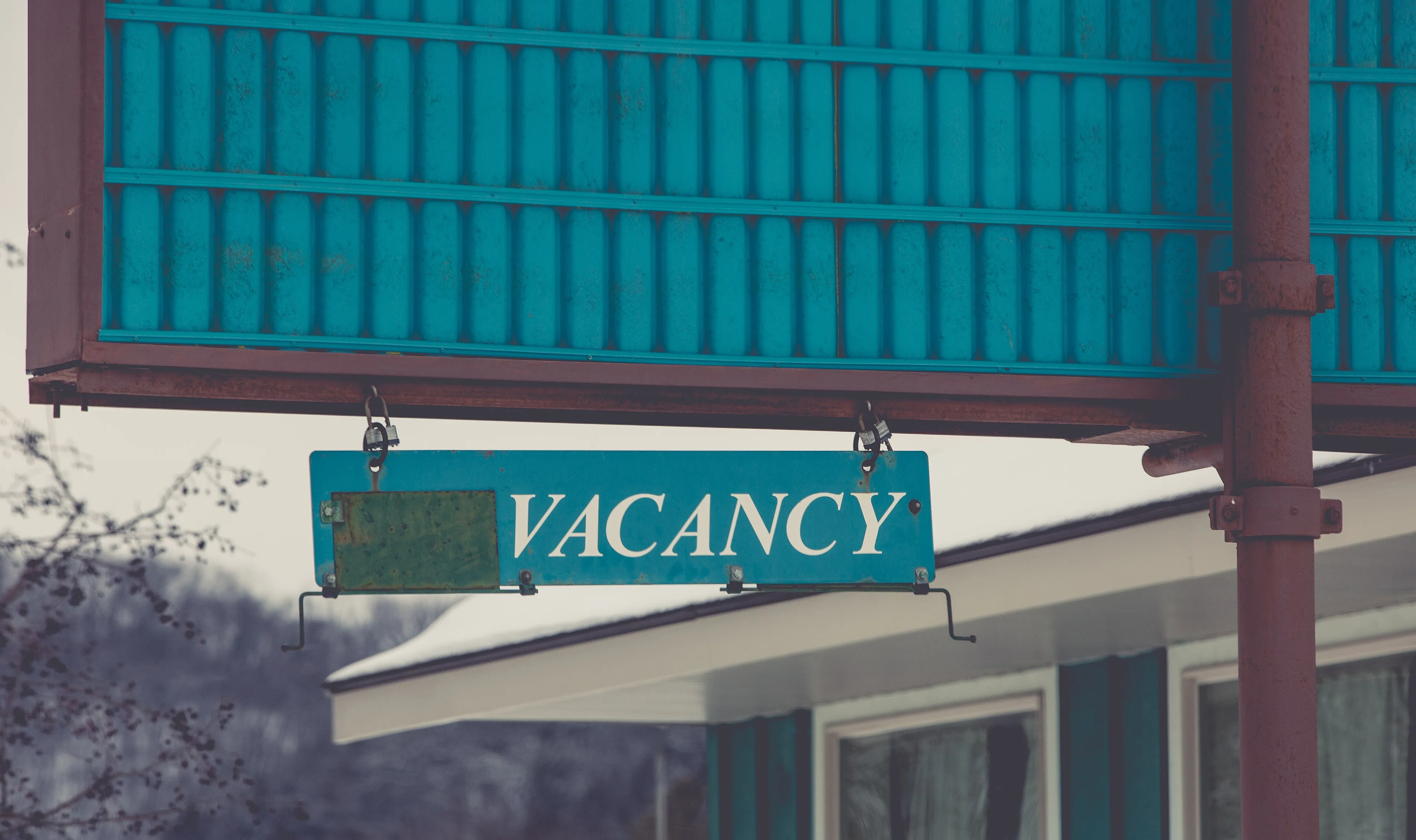 A white-on-teal VACANCY sign hanging under a blank motel sign box, with the motel roofline behind it