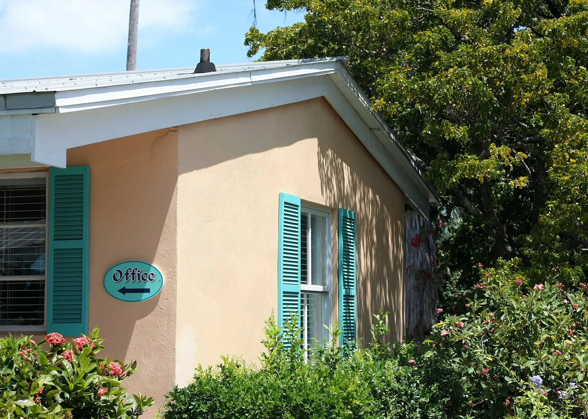 A peach-colored motel office corner with teal shutters and an oval Office sign with a directional arrow, framed by flowering shrubs