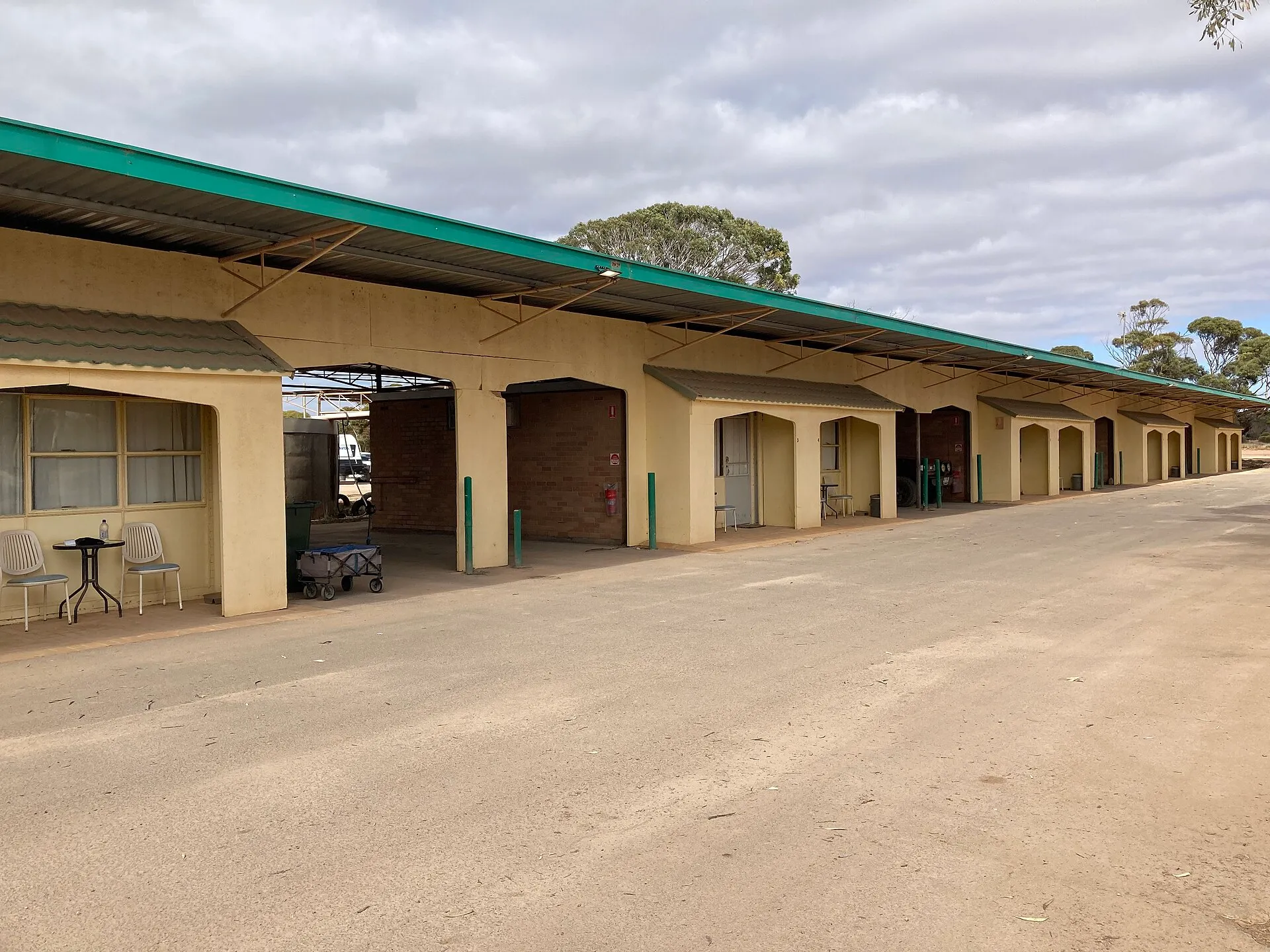 A long single-storey row of motel rooms under a green roof, with arched carports and two chairs outside a door