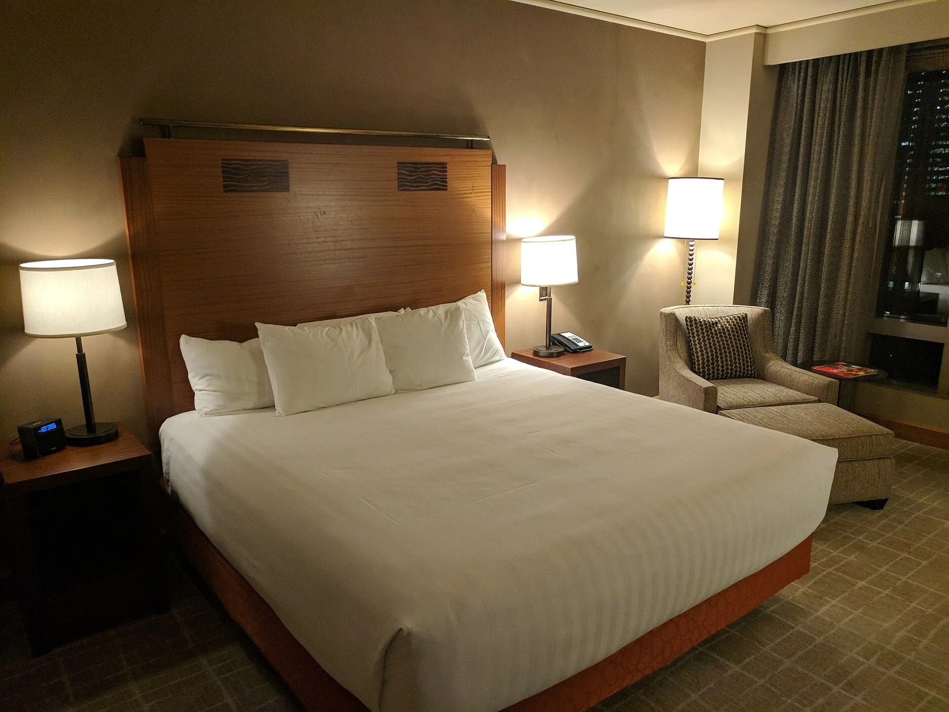 Hotel room at night with a made bed, two lit bedside lamps, a desk phone and an armchair by a curtained window