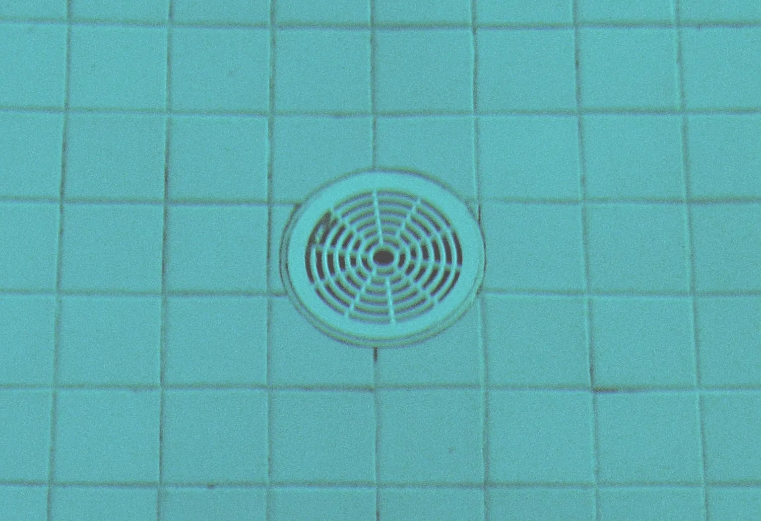 A round slotted drain cover set in teal tile on the floor of a swimming pool